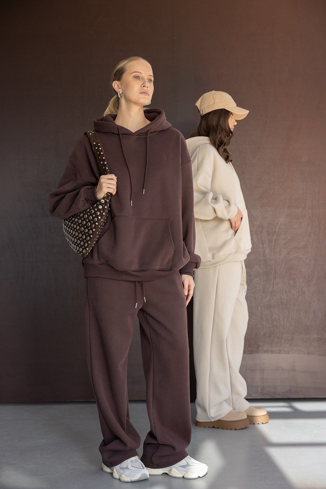 שתי נשים לובשות סט טרנינג רחב ואופנתי בצבע חום ובז', אחת עם תיק צד, רקע ניטרלי, אופנת נשים קז'ואל