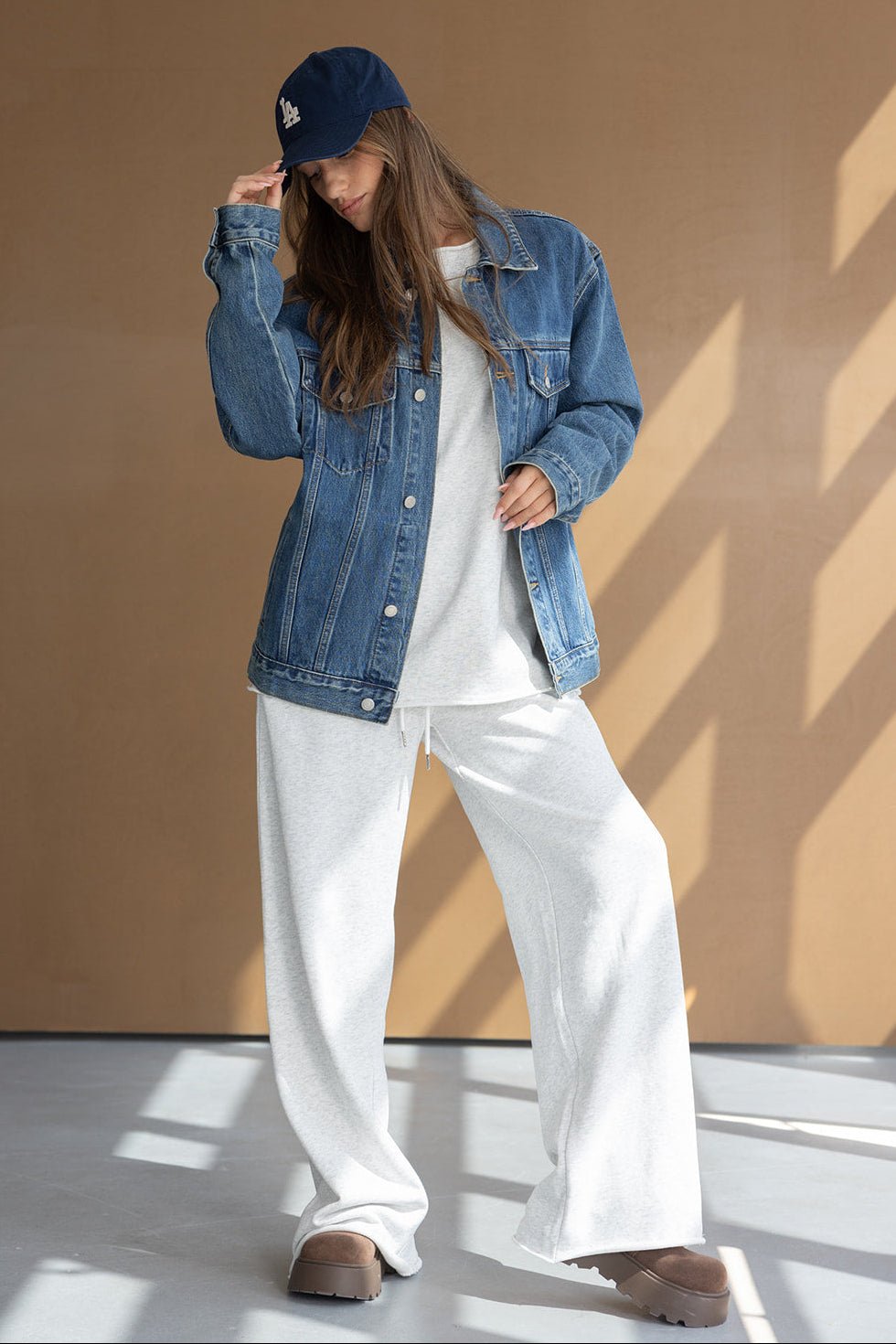 אישה עם ז'קט ג'ינס מעל חולצה אפורה רחבה, מכנסי טרנינג לבנים, כובע כחול ונעלי פלטפורמה חומות, לוק קז'ואל אופנתי לנשים