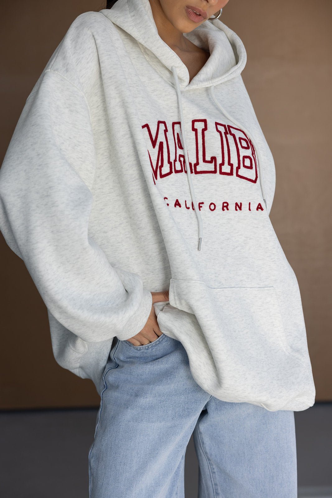 אישה לובשת קפוצ׳ון אוברסייז אפור עם כיתוב MALIBU אדום ומכנסי ג׳ינס, אופנה לנשים