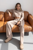 דוגמנית בלוק קז'ואל אופנתי יושבת על ספת עור, לובשת סט בגדי פנאי בצבע בז' ומוקסינים תואמים