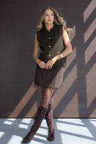 דוגמנית לובשת וסט שחור עם כפתורים, חצאית קצרה כהה ומגפיים גבוהים לאופנת נשים צעירות