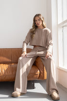 אישה לובשת סט טרנינג בז' עם מכנסיים רחבים וסווטשירט קרופ, יושבת על ספה חומה ליד חלון, אופנה לנשים