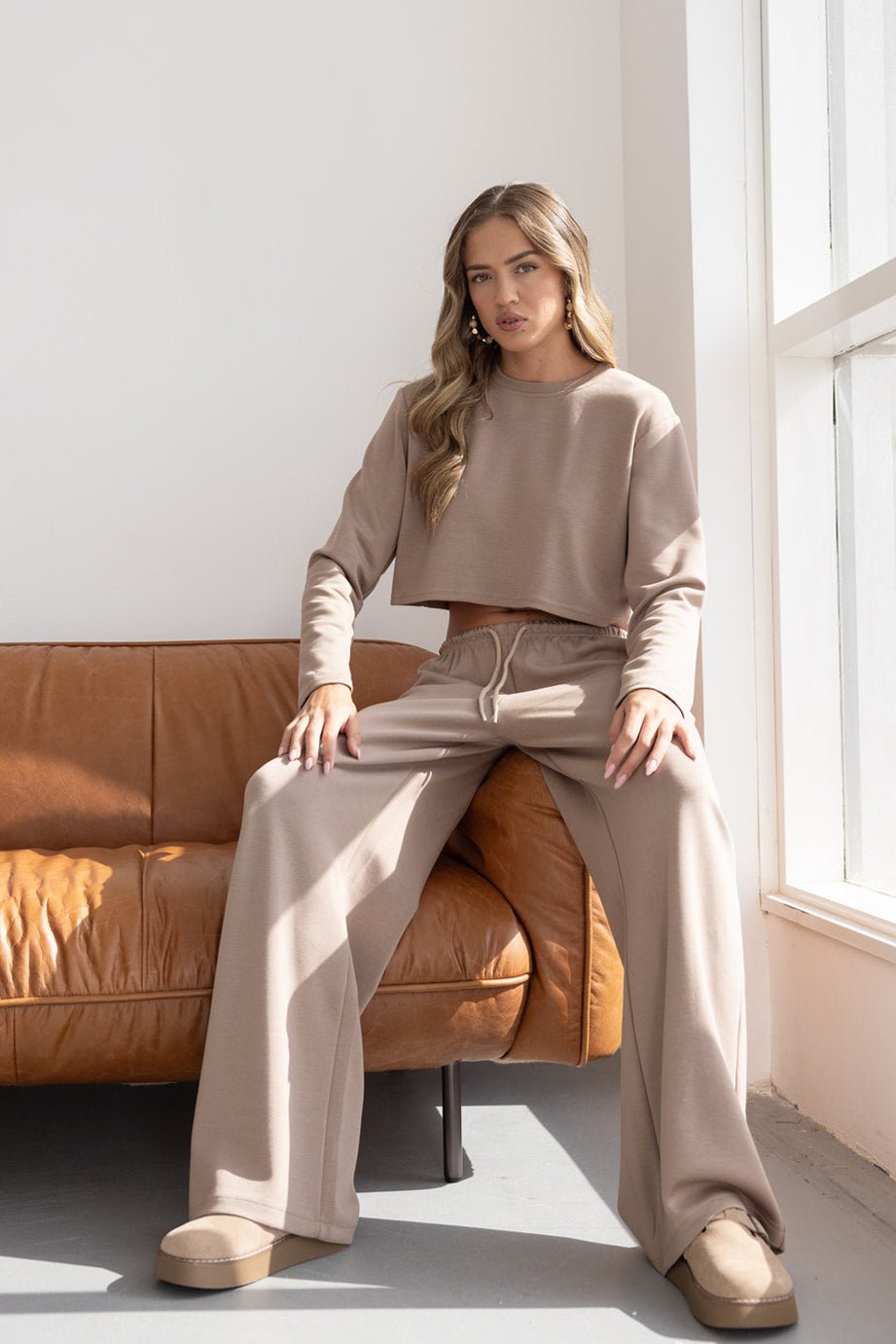 אישה לובשת סט טרנינג בז' עם מכנסיים רחבים וסווטשירט קרופ, יושבת על ספה חומה ליד חלון, אופנה לנשים