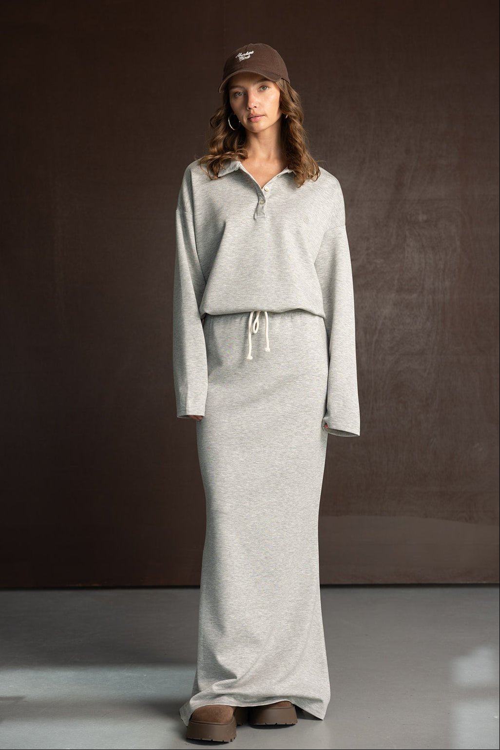 אישה דוגמנית לובשת שמלת סווטשירט אפורה ארוכה עם שרוך, כובע חום ונעלי פלטפורמה חומות, רקע חום