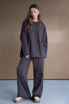 אישה לובשת סט טרנינג אופנתי בצבע אפור כהה, חולצה ארוכה ומכנסיים רחבים, עם נעליים פלטרפורמה, רקע ניטרלי