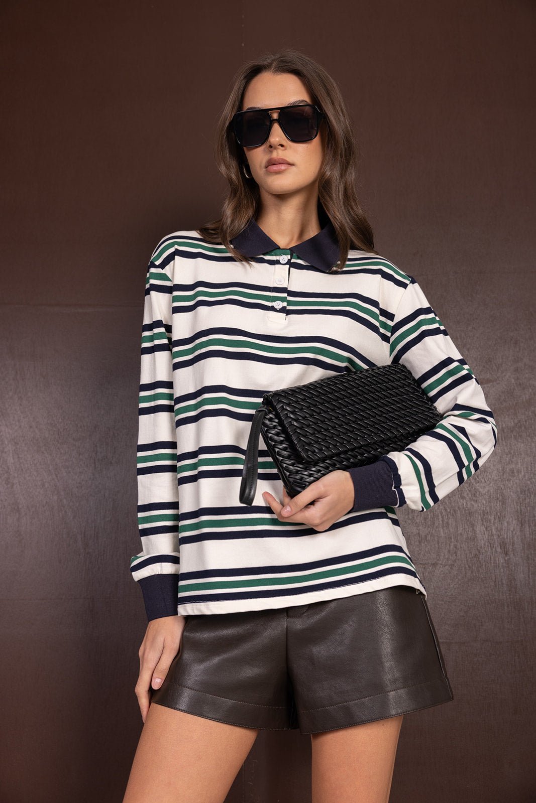 אישה עם חולצת פולו פסים, מכנסי עור קצרים, תיק קלאץ' קלוע ומשקפי שמש אופנתיים