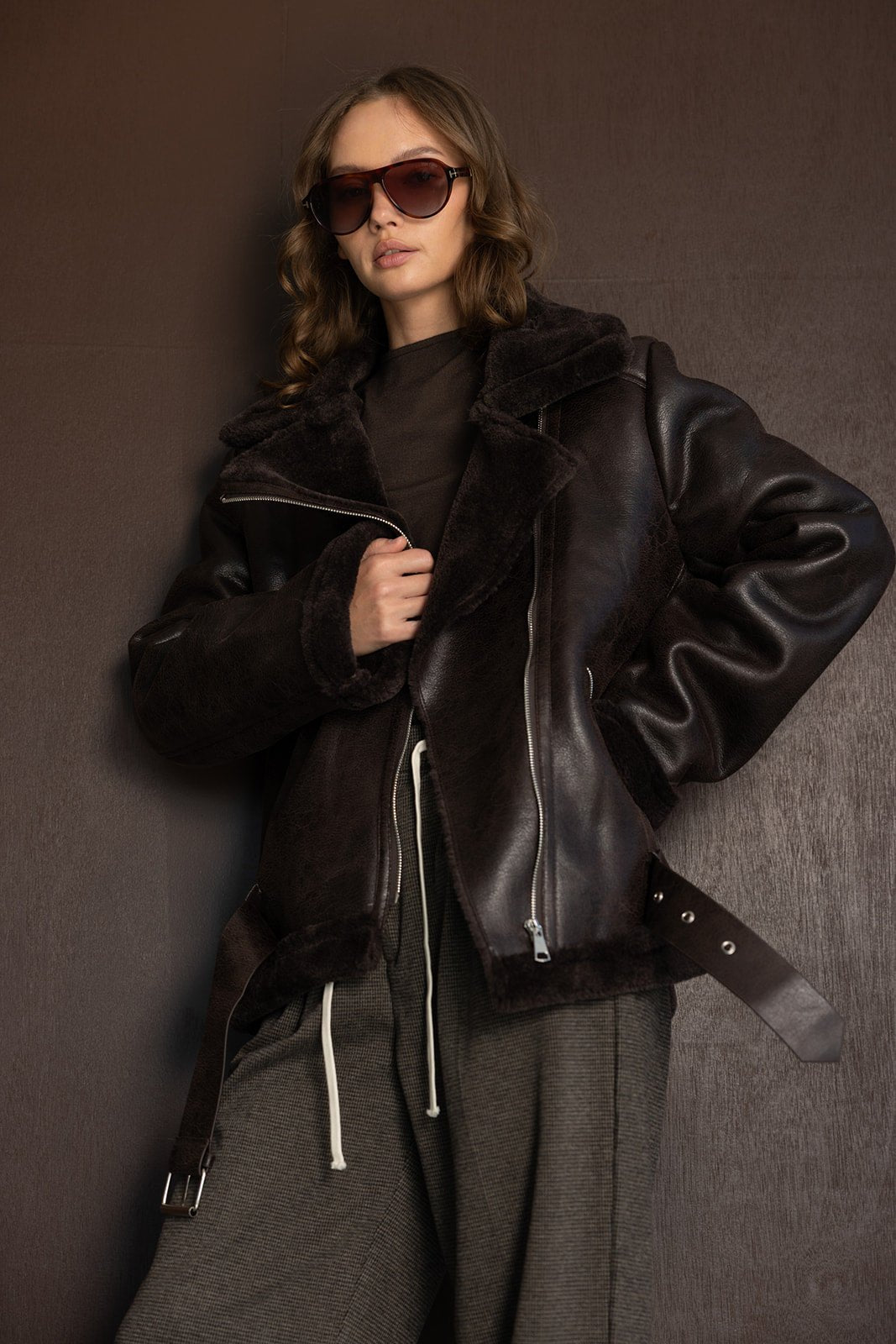 דוגמנית לובשת מעיל עור חום אופנתי עם רוכסן, משקפי שמש וחולצה כהה, אופנת חורף לנשים