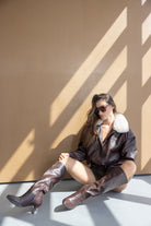 דוגמנית עם מעיל עור חום עם צווארון פרווה, מגפיים חומים ומשקפי שמש, יושבת בשמש