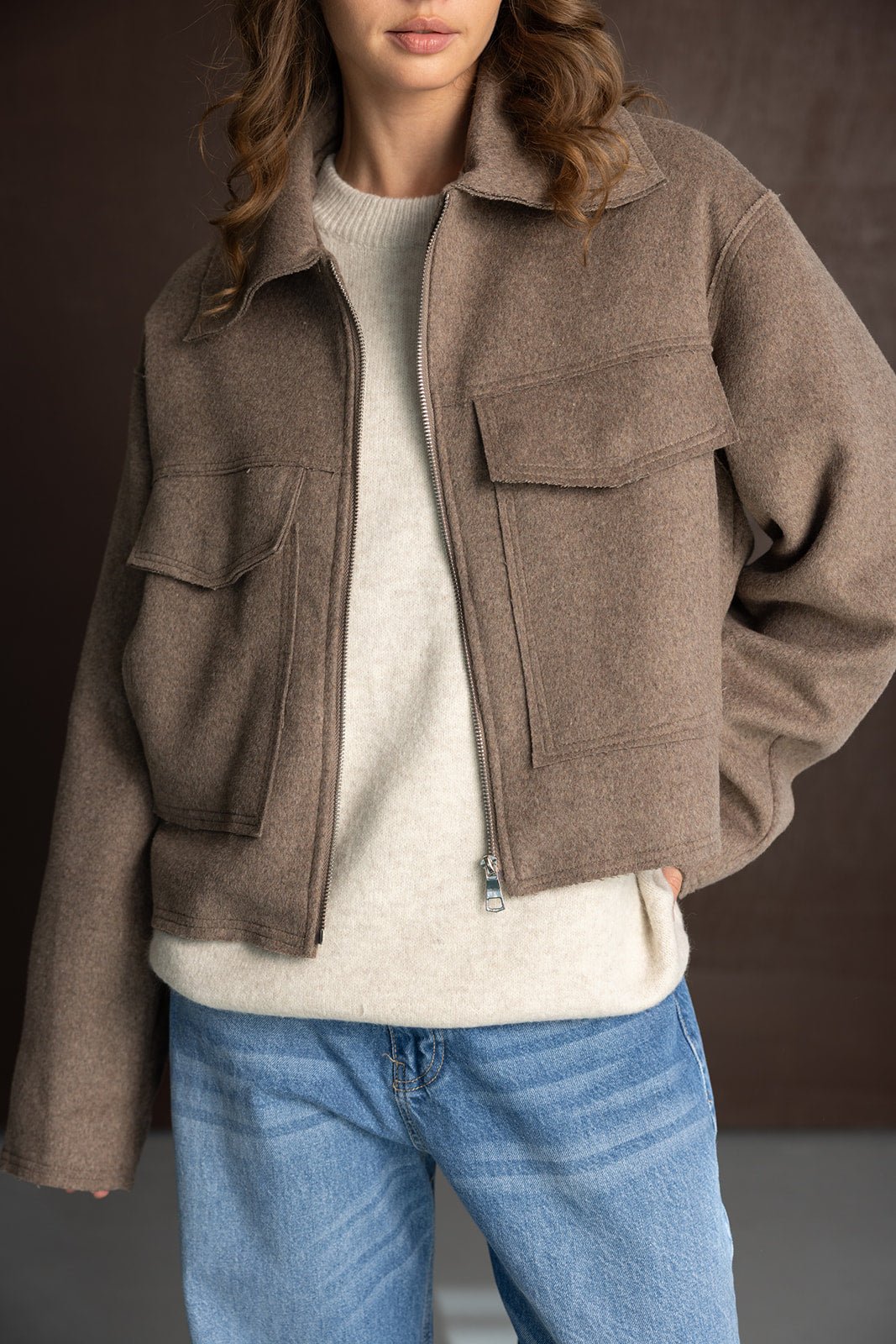 דוגמנית לובשת ג'קט חום קצר עם כיסים, סוודר קרם ומכנסי ג'ינס לנשים