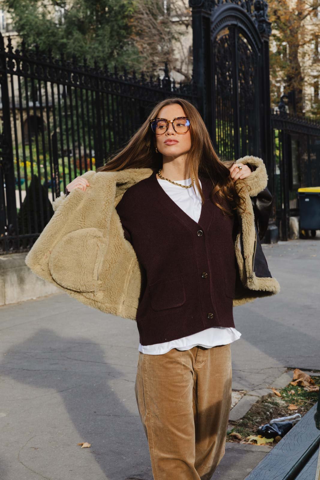 אישה עם משקפיים בלבוש חורפי אופנתי, ג'קט פרוותי פתוח, סוודר בורדו, מכנסי קורדרוי בז' ברחוב עירוני