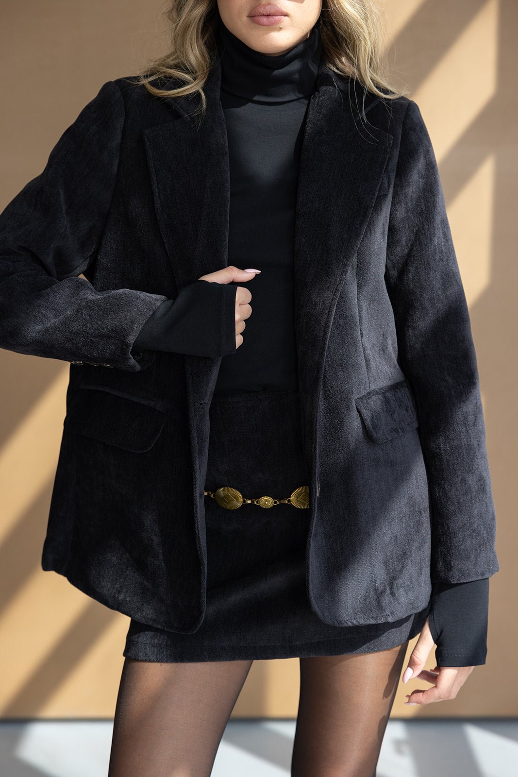 אישה לובשת בלייזר שחור עם חצאית מיני, טופ גולף שחור וגרביון שקוף, חגורת זהב אופנתית