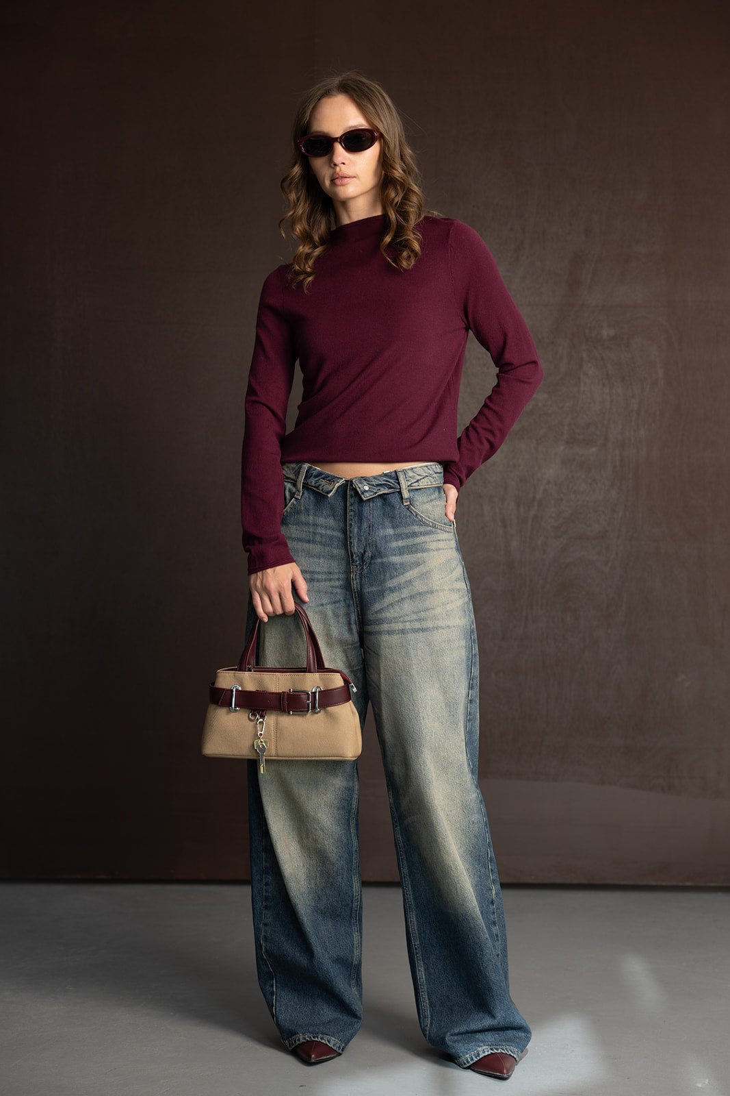 אופנת נשים צעירה: דוגמנית עם ג'ינס רחב, סוודר בורדו ומשקפי שמש, מחזיקה תיק בז'