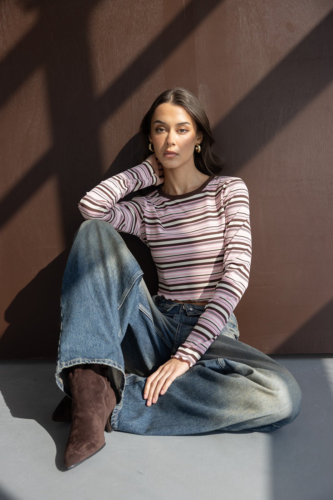 אישה עם חולצה מפוספסת ארוכת שרוולים בגווני ורוד וחום, ג'ינס רחב ומגפיים חומים יושבת על הרצפה