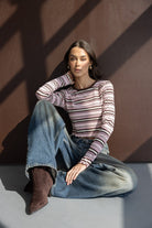 אישה עם חולצה מפוספסת ארוכת שרוולים בגווני ורוד וחום, ג'ינס רחב ומגפיים חומים יושבת על הרצפה