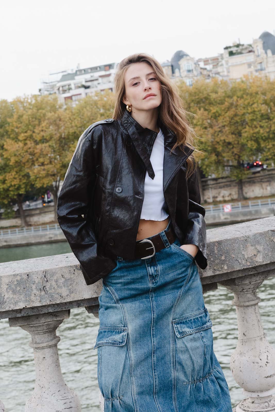 אישה לובשת ז'קט עור שחור, חולצה לבנה קצרה וחצאית ג'ינס, עומדת ליד נהר בסביבה עירונית