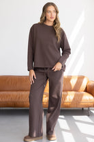 אישה לובשת סט טרנינג חום רחב, נעלי בית, עומדת בסלון מודרני על ספה בצבע חום