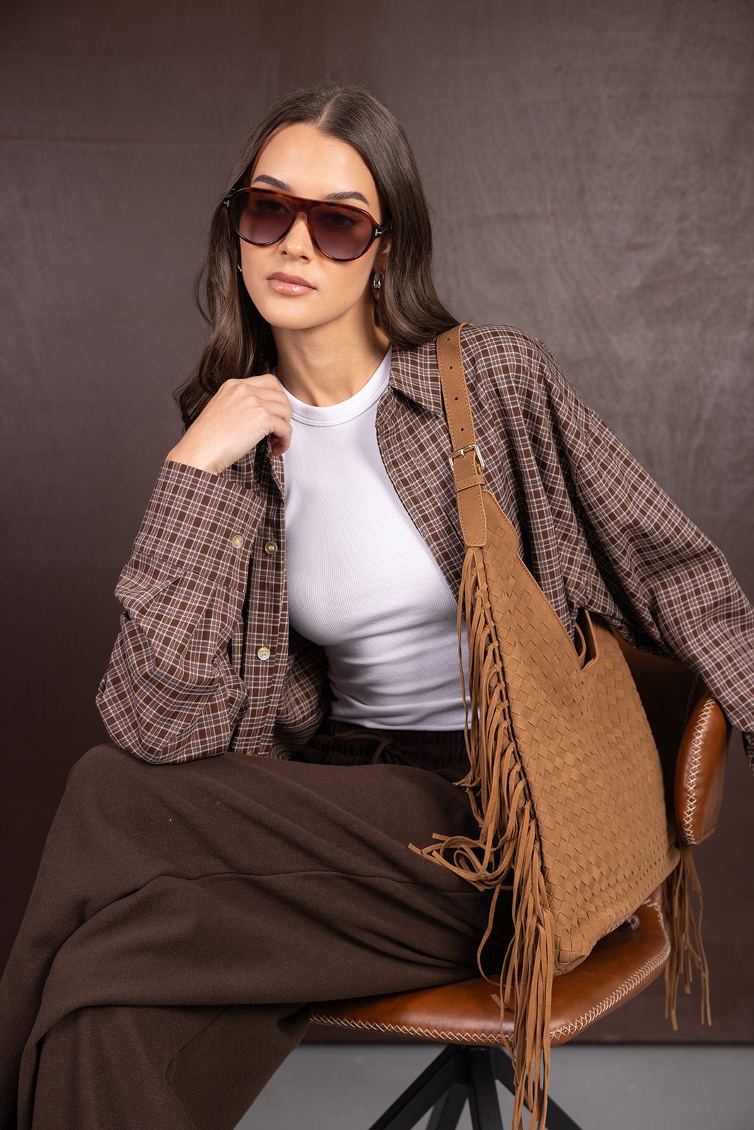 אישה עם חולצה משובצת חומה, גופיה לבנה, מכנסיים חומים, תיק קש חום ומשקפי שמש אופנתיים