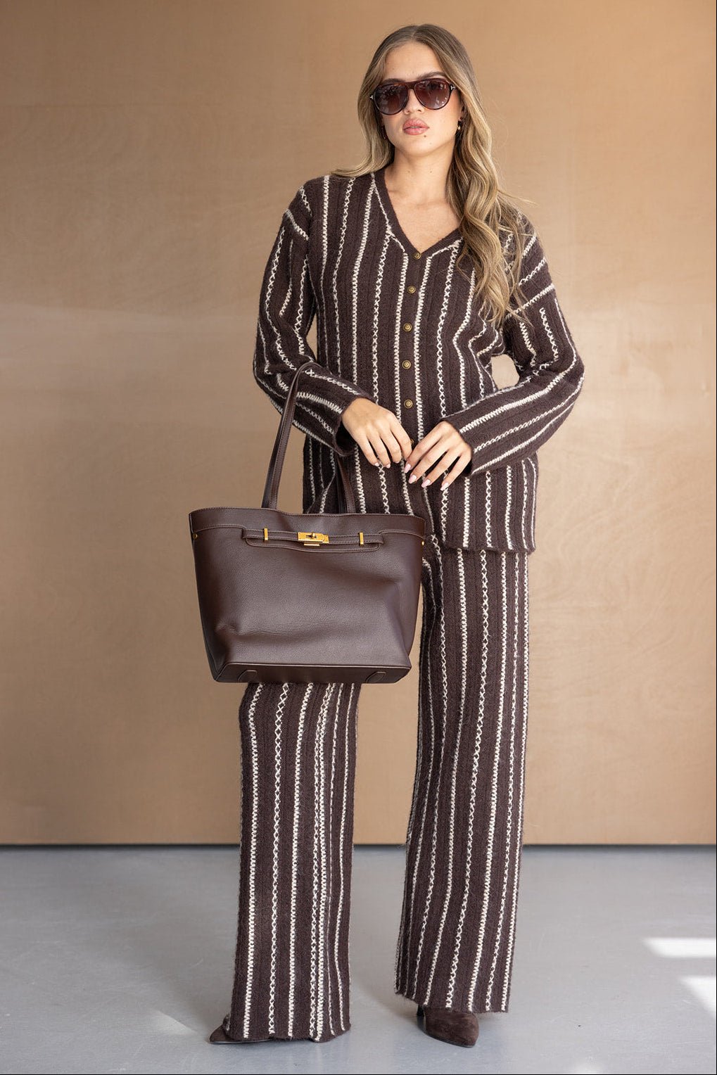אישה עם חליפת פסים חומה ולבנה, תיק יד חום ומשקפי שמש, אופנת נשים חורפית