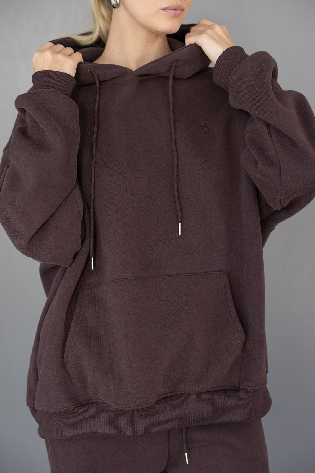 אישה לובשת קפוצ'ון חום כהה במראה אופנתי עם כיס קדמי ושרוכים, רקע אפור