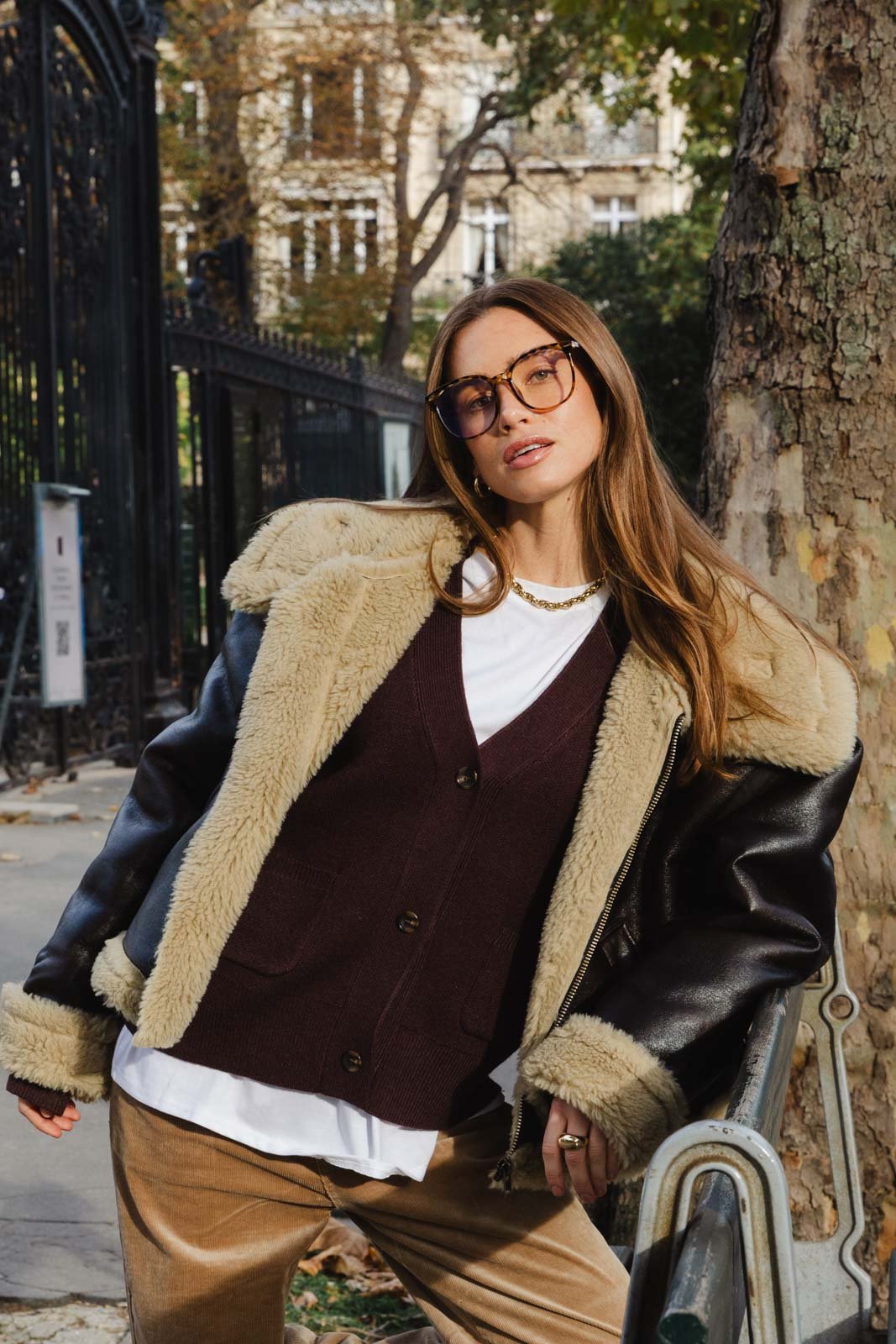 אישה על ספסל ברחוב לובשת מעיל עור עם פרווה, סריג חום ומשקפי אופנה, אופנה חורפית לנשים