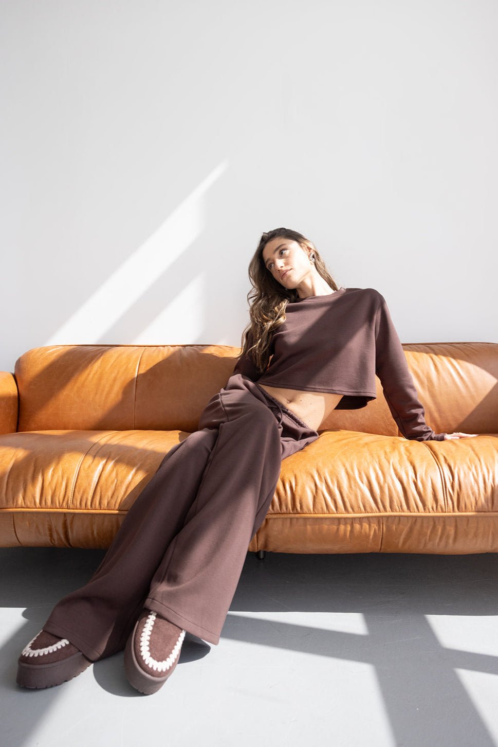 אישה בלבוש סט טרנינג חום יושבת על ספה עור, אופנת נשים מודרנית וסטייל צעיר