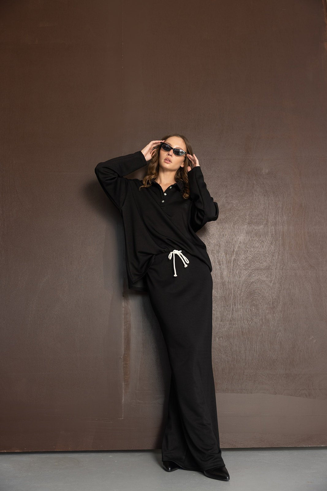 דוגמנית לובשת סט טרנינג שחור בסגנון קז'ואל עם חולצה שרוול ארוך, חצאית ומכנסיים, משקפי שמש אופנתיים, רקע חום