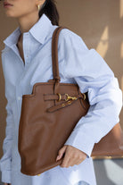 אישה עם חולצה מכופתרת תכלת-פסים נושאת תיק כתף חום אופנתי עם אבזמים זהובים