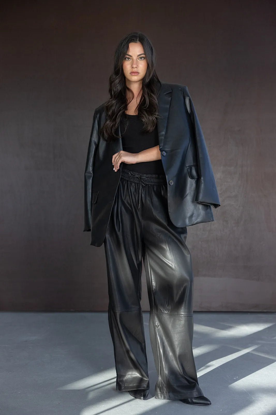 אישה עם שיער כהה לובשת חליפה שחורה דמוי עור, מכנסיים רחבים וז'קט על כתפיה, אופנת נשים עדכנית