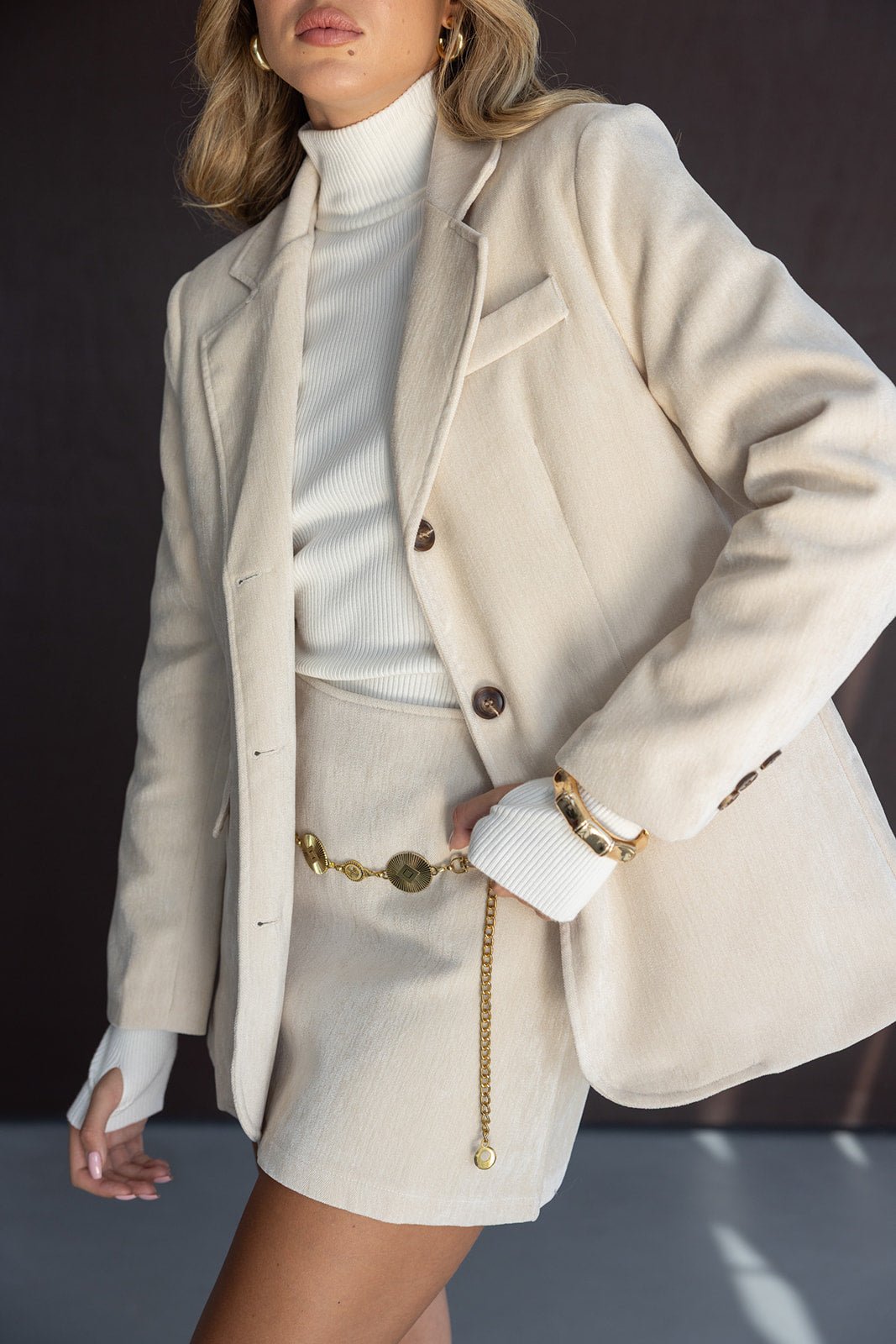 אישה לובשת בלייזר וחצאית בצבע בז', סוודר לבן וגולף, עם חגורת שרשרת זהב, אופנת נשים אלגנטית