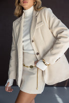 אישה לובשת בלייזר וחצאית בצבע בז', סוודר לבן וגולף, עם חגורת שרשרת זהב, אופנת נשים אלגנטית