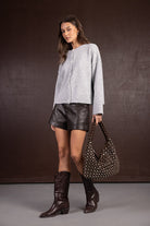 דוגמנית לובשת קרדיגן אפור, מכנסי עור קצרים שחורים, מגפיים חומים ותיק כתף עם ניטים, אופנה לנשים