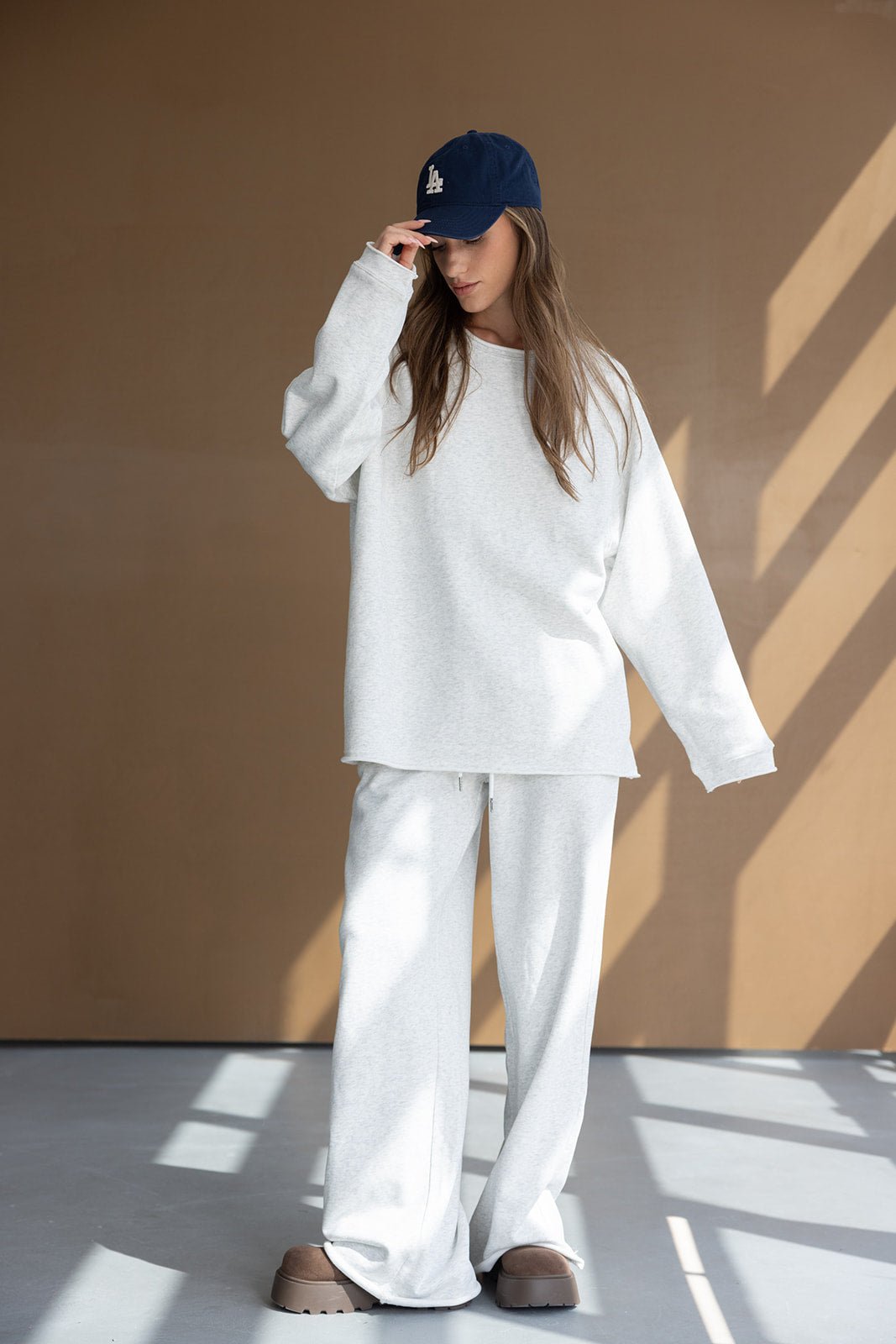 אישה לובשת סווטשירט ומכנסיים אפורים רחבים, כובע כחול ונעלי בית חומות, לוק קז'ואל אופנתי.