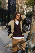 אישה לובשת מעיל עור שחור עם פרווה, קרדיגן בורדו ומכנסיים בגוון קאמל, עומדת ברחוב עירוני