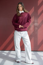 אישה עם סוודר בורדו, חולצה לבנה מתחת וג'ינס רחב אופנתי, עומדת על רקע קיר אדום