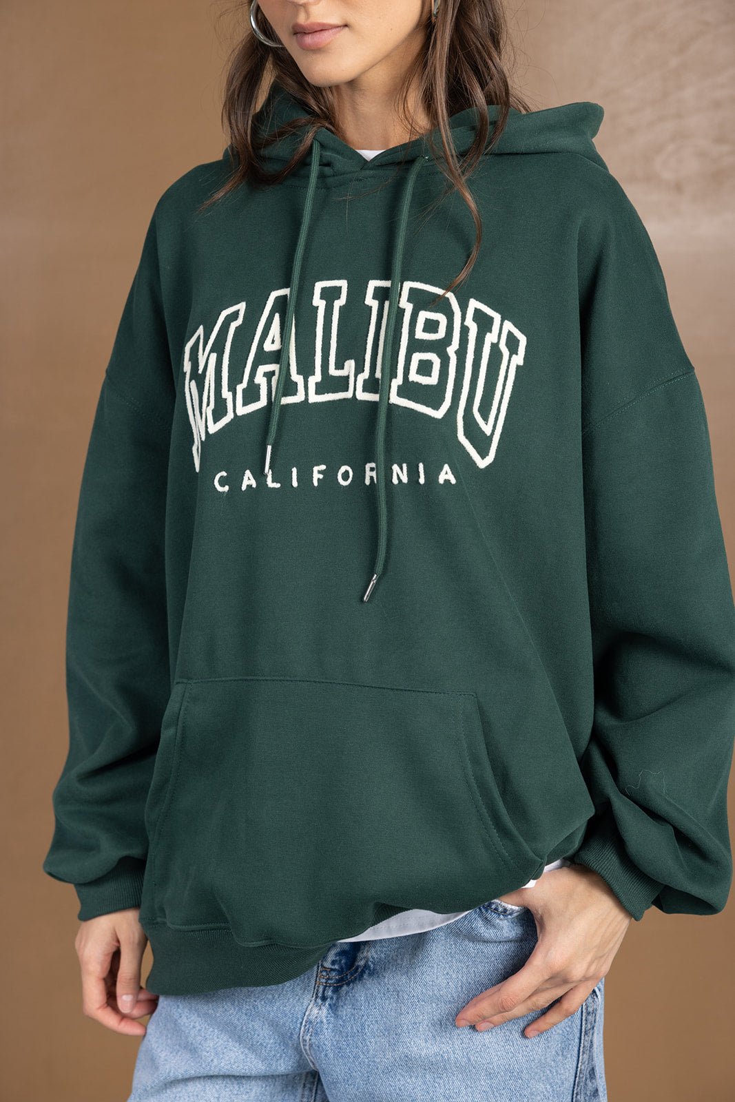 אישה לובשת קפוצ׳ון ירוק כהה עם כיתוב MALIBU CALIFORNIA וג׳ינס אופנתי