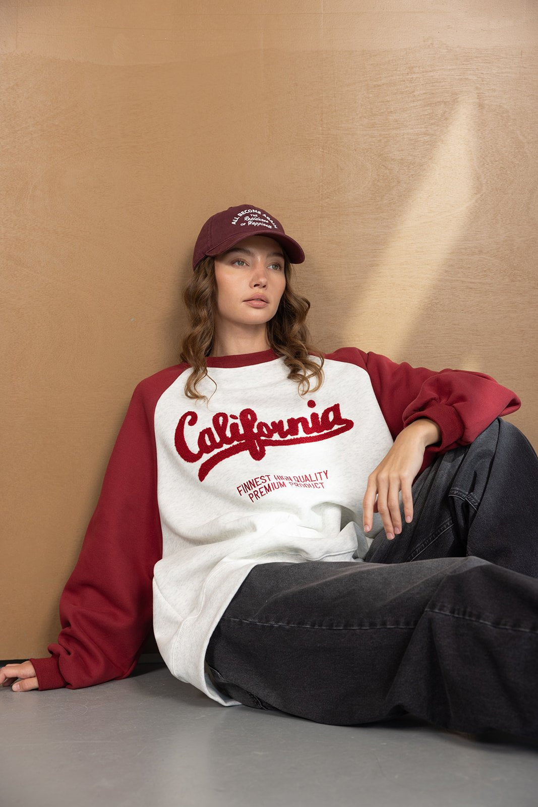 דוגמנית לובשת סווטשירט נשים אופנתי עם כיתוב California, שרוולים בורדו, וכובע תואם, יושבת ברקע נייטרלי