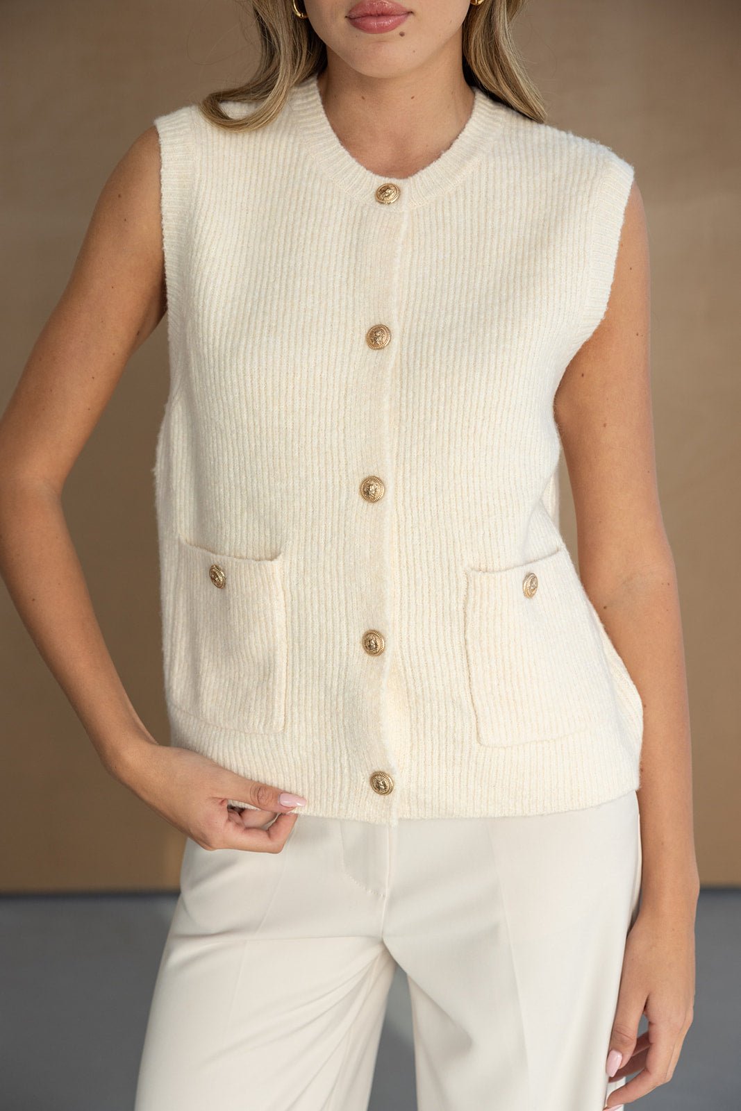 אישה לובשת וסט סריג שמנת ללא שרוולים עם כפתורים זהובים וכיסים, אופנת נשים אלגנטית