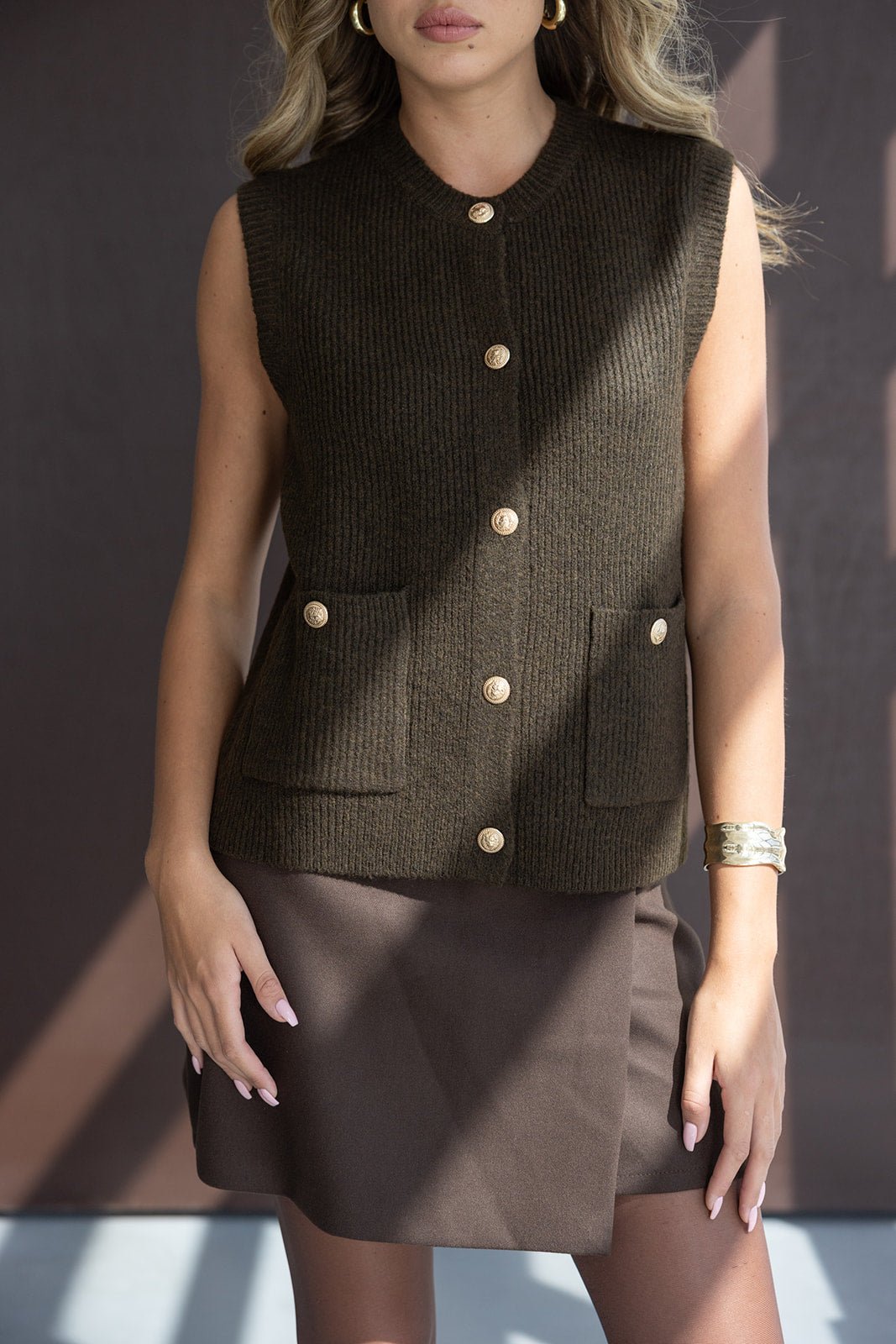 אישה לובשת וסט סרוג חום עם כפתורים זהובים וחצאית חומה, אופנה לנשים צעירות