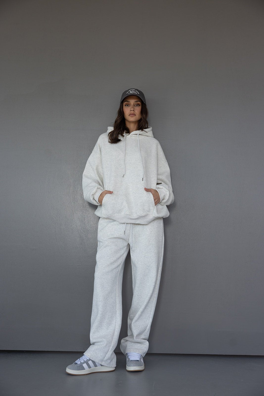 אישה מדגמנת סט טרנינג נשים אפור עם קפוצ'ון, מכנסיים רחבים וכובע בצבע כהה, לוק קז'ואל אופנתי