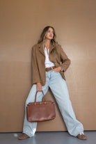 אישה לבושה בז'קט חום, חולצה לבנה ומכנסי ג'ינס רחבים, מחזיקה תיק חום אופנתי