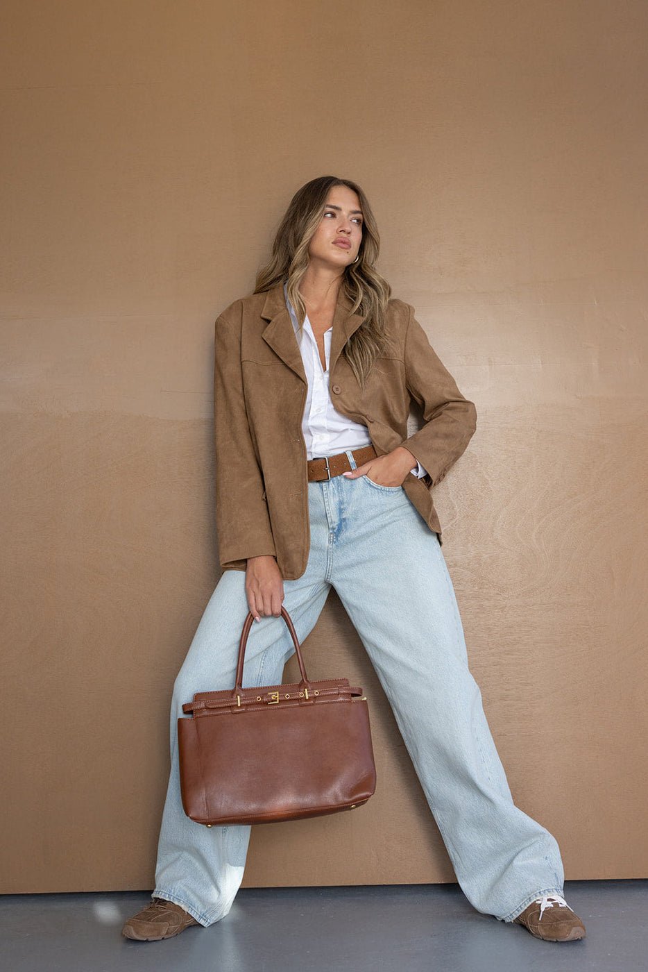 אישה לבושה בז'קט חום, חולצה לבנה ומכנסי ג'ינס רחבים, מחזיקה תיק חום אופנתי