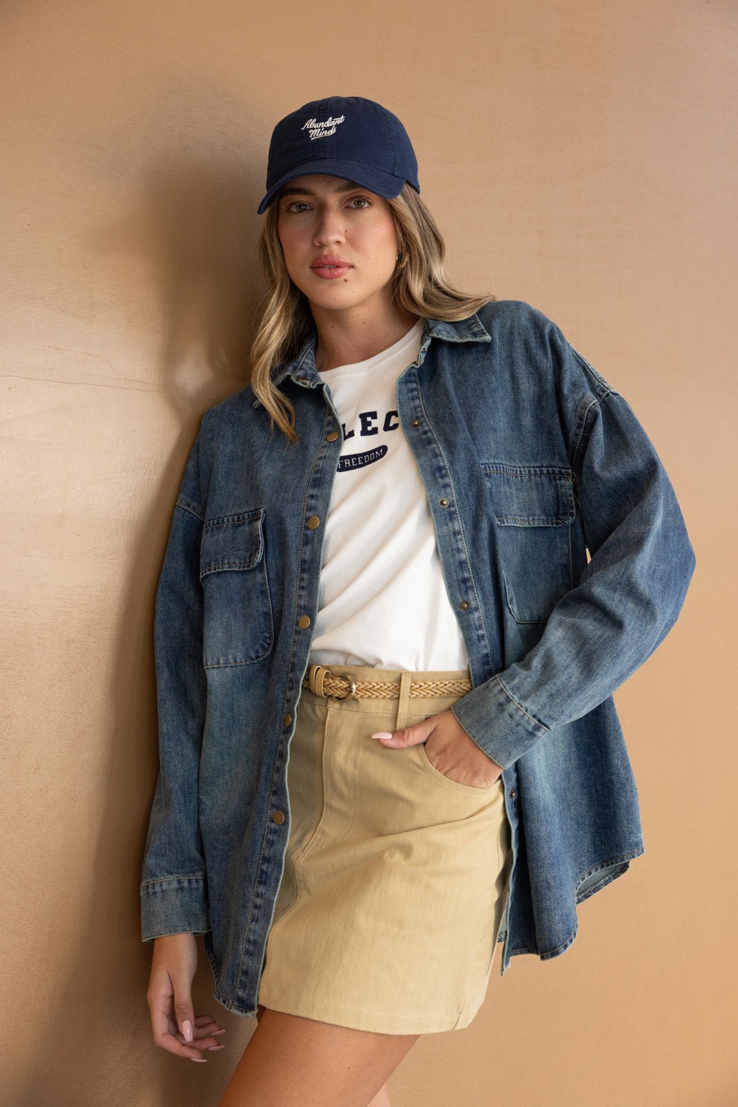 דוגמנית צעירה לובשת חולצת ג'ינס אוברסייז, חצאית בז', טי-שירט וכובע כחול, לוק קז'ואל לנשים