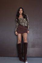 אישה לובשת בגד גוף מנומר, חצאית חומה קצרה ומגפיים חומים על רקע חום, אופנת נשים צעירה