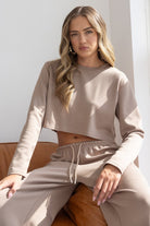 דוגמנית יושבת על ספה עם סט טרנינג נשים בצבע בז', חולצה קרופ ומכנסיים ארוכים לעצמאות