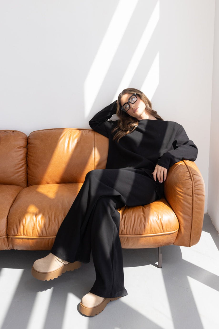 אשה אופנתית יושבת על ספה חומה עם חליפה שחורה ונעלי פלטפורמה בהירות, משקפי שמש, סטודיו מואר