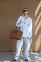 אישה לובשת חולצה מכופתרת פסים כחולים, ג'ינס רחב, נעליים חומות ותיק עור חום, לוק אופנתי לנשים