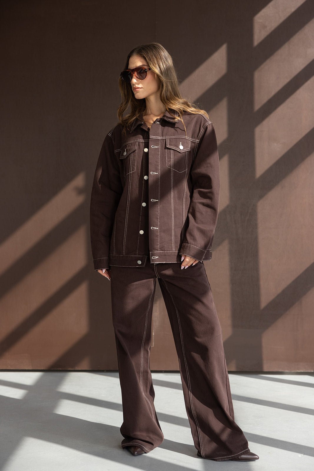דוגמנית לובשת ג'קט ומכנסיים חומים בגזרה רחבה עם תפרים בהירים ומרכיבה משקפי שמש, אופנת נשים עדכנית