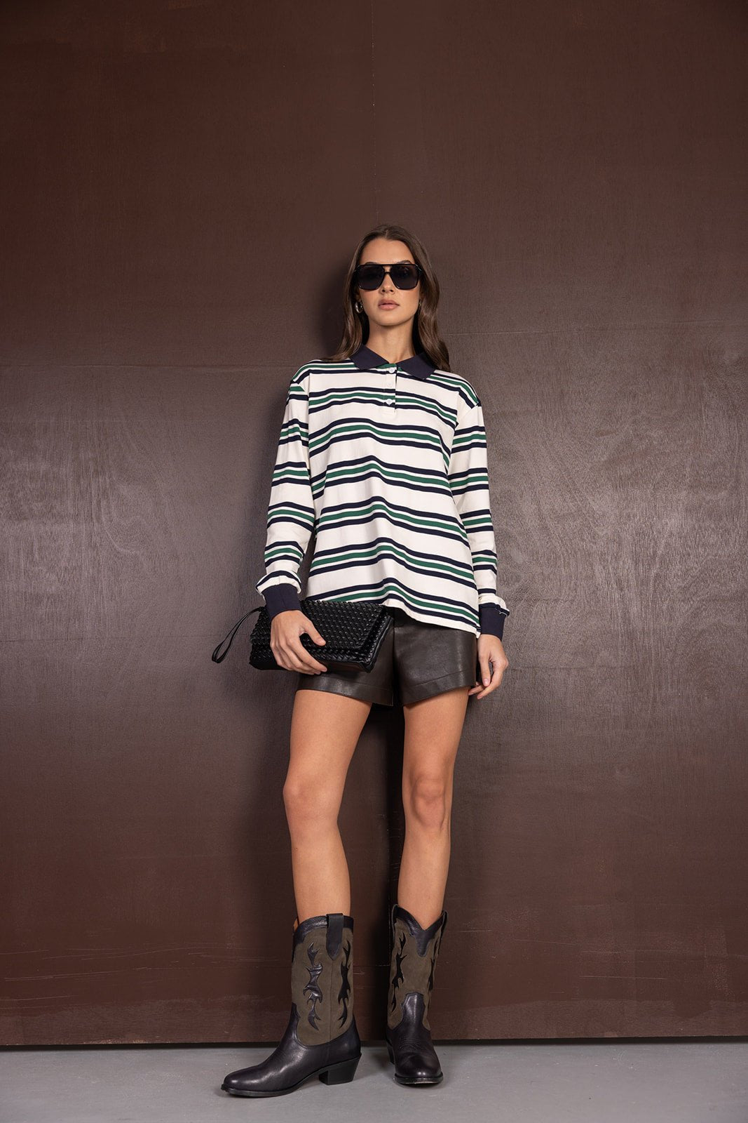 אישה עם חולצת פסים ארוכה, מכנסי עור קצרים ומגפיים אופנתיים, אוחזת תיק ערב