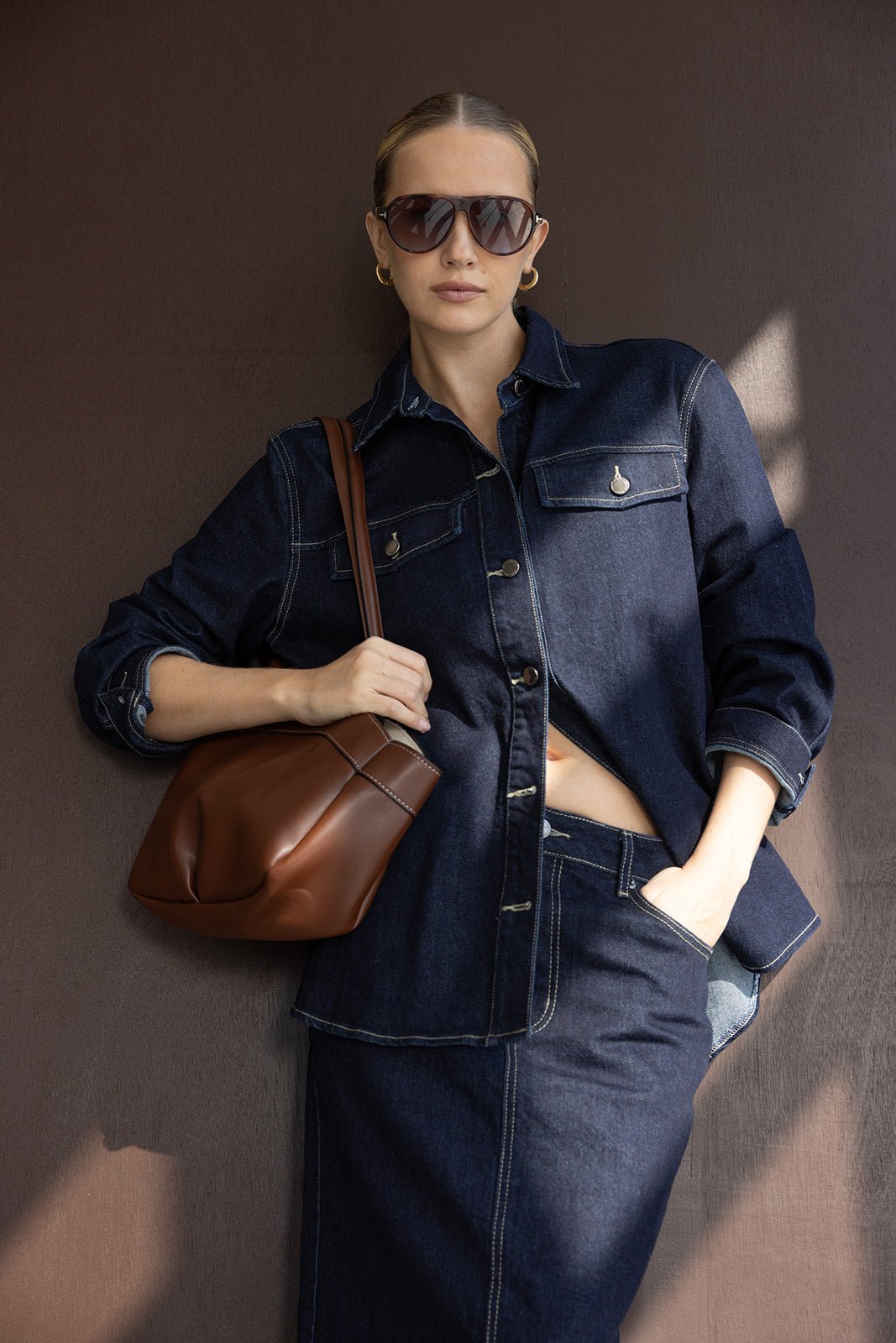 דוגמנית לובשת חולצת ג'ינס כהה עם חצאית תואמת, מרכיבה משקפי שמש ומחזיקה תיק חום, אופנת נשים עדכנית