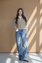 אישה עם שיער ארוך לובשת סוודר פסים חום-לבן, ג'ינס ישר ומשקפי שמש, עומדת ברקע חום מואר